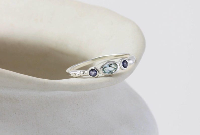 May include: A silver ring with three oval-shaped gemstones. The center stone is a light blue aquamarine, and the two smaller stones on either side are purple.