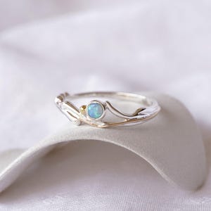 May include: A silver ring with a light blue opal gemstone. The ring has a delicate, nature-inspired design with gold accents. The opal is round and set in a bezel setting. The ring is displayed on a white surface.