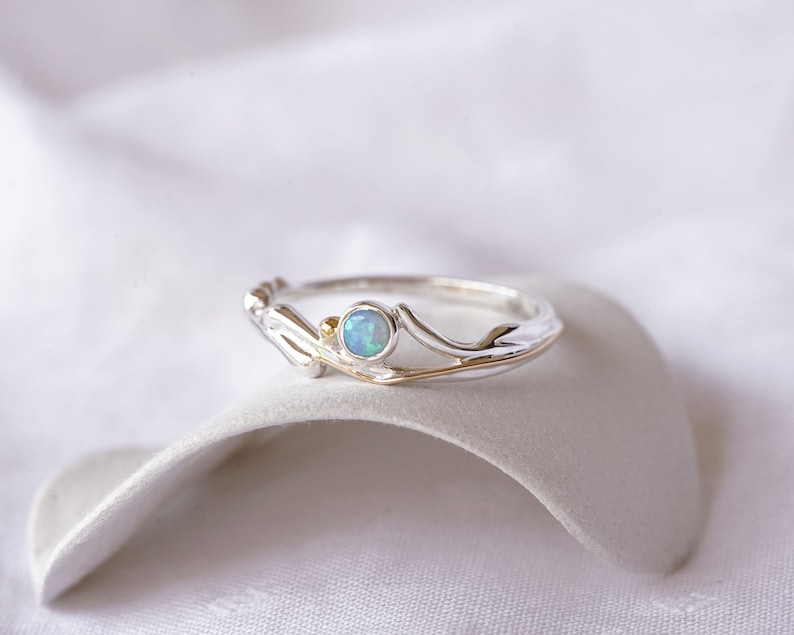 May include: A silver ring with a light blue opal gemstone. The ring has a delicate, nature-inspired design with gold accents. The opal is round and set in a bezel setting. The ring is displayed on a white surface.
