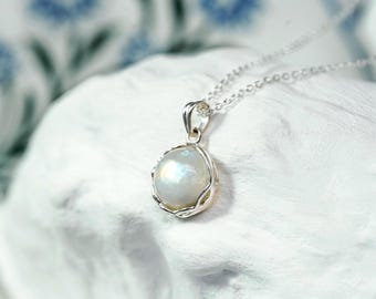 May include: A silver necklace with a pearlescent, oval-shaped pendant. The pendant is set in a decorative silver frame and hangs from a delicate silver chain. The background is a textured white surface.