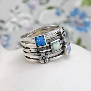 May include: A collection of silver rings featuring square, blue opal gemstones. The rings have a layered design with a flower and spiral detail. The rings are on a white surface with a floral background.