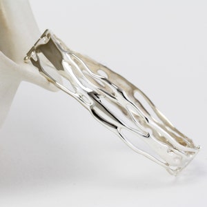 May include: A silver-colored openwork cuff bracelet. The bracelet has an organic, flowing design with irregular, interconnected lines. The jewelry is displayed against a white background, highlighting its reflective surface and intricate details. The bracelet's design suggests a modern, artistic aesthetic.