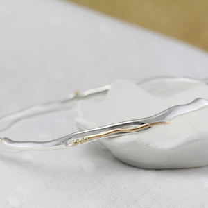 May include: Two silver bangles with a wavy design, accented with gold-coloured details. The bracelets are displayed on a white, textured surface, showcasing their elegant and organic form. The jewellery is suitable for everyday wear.