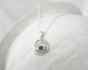 May include: A silver pendant necklace featuring a round, faceted green gemstone set in a circular frame. The frame is composed of intertwined silver and thin gold-toned wire accents, suspended from a delicate silver chain with a fine link pattern.