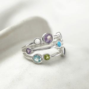 May include: A silver ring featuring three bands, each embellished with various coloured gemstones. The gemstones include purple, blue, green, and white, creating a vibrant design. The ring is set against a soft, white background.