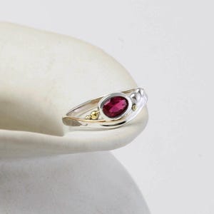 May include: A silver ring with a red gemstone and gold accents. The ring is set on a white surface.