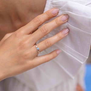 May include: A silver ring with a light blue gemstone, set in a delicate, nature-inspired design. The ring is on a person's finger, with a white garment in the background. The gemstone is round and appears to be an opal.