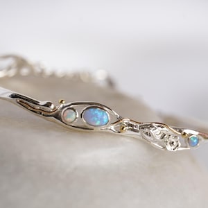 Può includere: Un braccialetto rigido in argento con un design delicato e organico con tre pietre di opale. Il braccialetto ha una forma ondulata e fluida ed è impreziosito da dettagli dorati.
