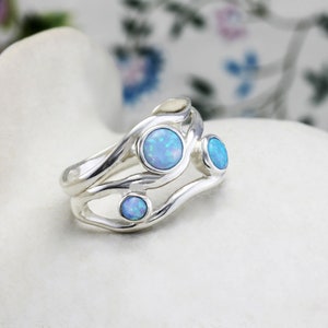 May include: A silver ring with a flowing design, featuring four round, light blue opal stones. The ring is set on a white surface, with a blurred floral background.