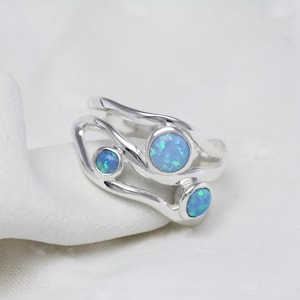 May include: A silver ring with three blue opal stones. The ring has a unique, flowing design with the opals set in bezel settings. The opals have a vibrant blue color, and the ring is displayed on a white surface.