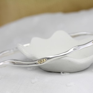 May include: Two silver bangles with a wavy design and gold accents rest on a white, petal-shaped dish. The bangles have a polished finish and a simple, elegant style. The background is a soft, blurred white and gold.