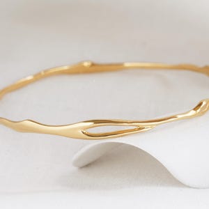 May include: A gold-coloured bangle bracelet with an organic, flowing design. The bracelet has an irregular shape with a smooth, polished finish. The jewellery is displayed on a white surface, creating a contrast with the gold.