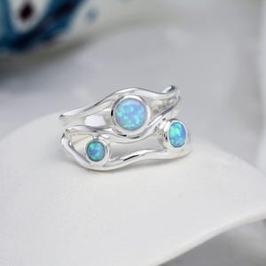 May include: Two silver rings with a flowing design, each featuring a light blue opal stone. The rings are stacked, showcasing the opals' iridescent quality. The rings are set against a white background.