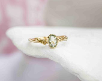 Puede incluir: Un anillo de oro con una piedra preciosa de color verde claro de corte ovalado engastada en un bisel. La banda del anillo tiene un diseño texturizado y orgánico que se asemeja a una rama de árbol fina con nudos sutiles.