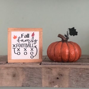 May include: A wooden sign with the text "Fall family FOOTBALL" and a football play diagram. The sign is next to a small orange pumpkin with a black stem and leaf.