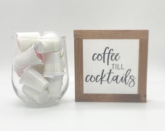 May include: A white wooden sign with the text "coffee till cocktails" in black lettering. A glass jar filled with white coffee pods is next to the sign.
