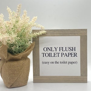 May include: A wooden sign with the text "ONLY FLUSH TOILET PAPER (easy on the toilet paper)" in black lettering on a white background. The sign is framed in a light brown wood frame.