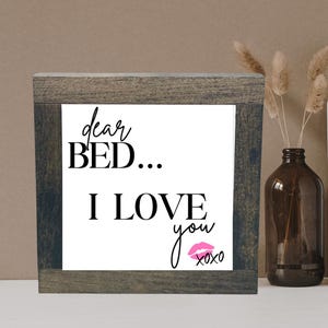 May include: A framed sign with the text "dear BED... I LOVE you xoxo" in black lettering on a white background. A pink lip print is below the text.