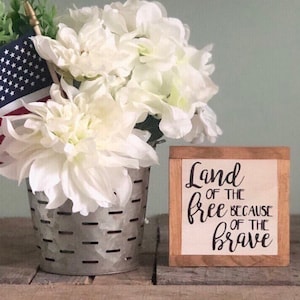 以下が含まれることがあります： 黒い書体で「Land of the free because of the brave」と書かれた木製の看板。看板は木製の表面に置かれ、金属製のバケツに白い花がアレンジされている。