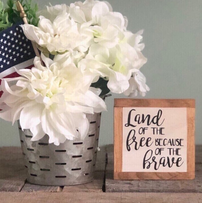 以下が含まれることがあります： 黒い書体で「Land of the free because of the brave」と書かれた木製の看板。看板は木製の表面に置かれ、金属製のバケツに白い花がアレンジされている。