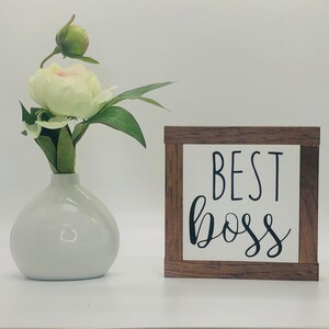 K&ouml;nnte beinhalten: Ein kleines Holzschild mit dem Text "BEST BOSS" in schwarzen Buchstaben auf wei&szlig;em Hintergrund. Das Schild steht auf einer wei&szlig;en Oberfl&auml;che neben einer wei&szlig;en Vase mit einer einzelnen wei&szlig;en Blume.