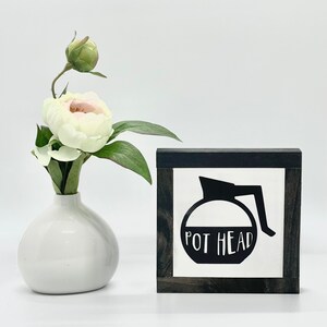 May include: A white wooden sign with a black frame and a black silhouette of a coffee pot with the text "POT HEAD" in white.