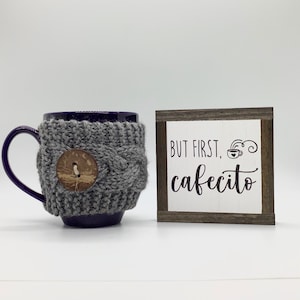 May include: A purple mug with a gray knitted cozy and a wooden button. The mug is next to a small wooden sign with the text "But first, cafecito" and a stylized coffee cup.