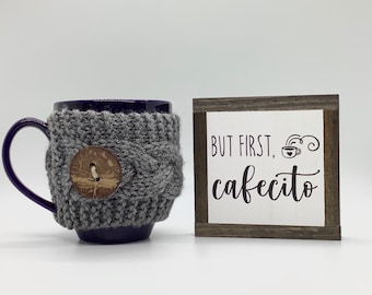 May include: A purple mug with a gray knitted cozy and a wooden button. The mug is next to a small wooden sign with the text "But first, cafecito" and a stylized coffee cup.