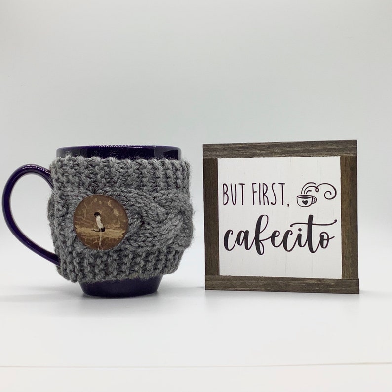 May include: A purple mug with a gray knitted cozy and a wooden button. The mug is next to a small wooden sign with the text "But first, cafecito" and a stylized coffee cup.