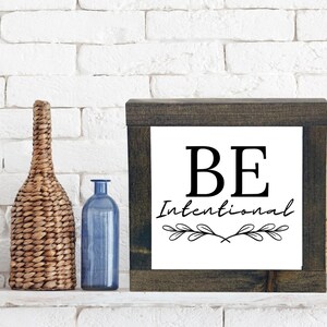 May include: A wooden framed sign with a white background and black text that reads "BE Intentional" with a decorative leaf design.