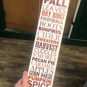 Puede incluir: Un letrero de madera blanco con las palabras "FALL" en letras grandes de color marr&oacute;n. Las palabras "LEAVES", "HAY RIDES", "FOOTBALL", "BOOTS", "BONFIRES", "CIDER", "SWEATERS", "HARVEST", "CUDDLES", "S'MORES", "PECAN PIE", "CARAMEL", "APPLES", "CORN MAZE", "PUMPKIN" y "SPICE" est&aacute;n listadas debajo en letras m&aacute;s peque&ntilde;as, marrones, naranjas y rosadas.