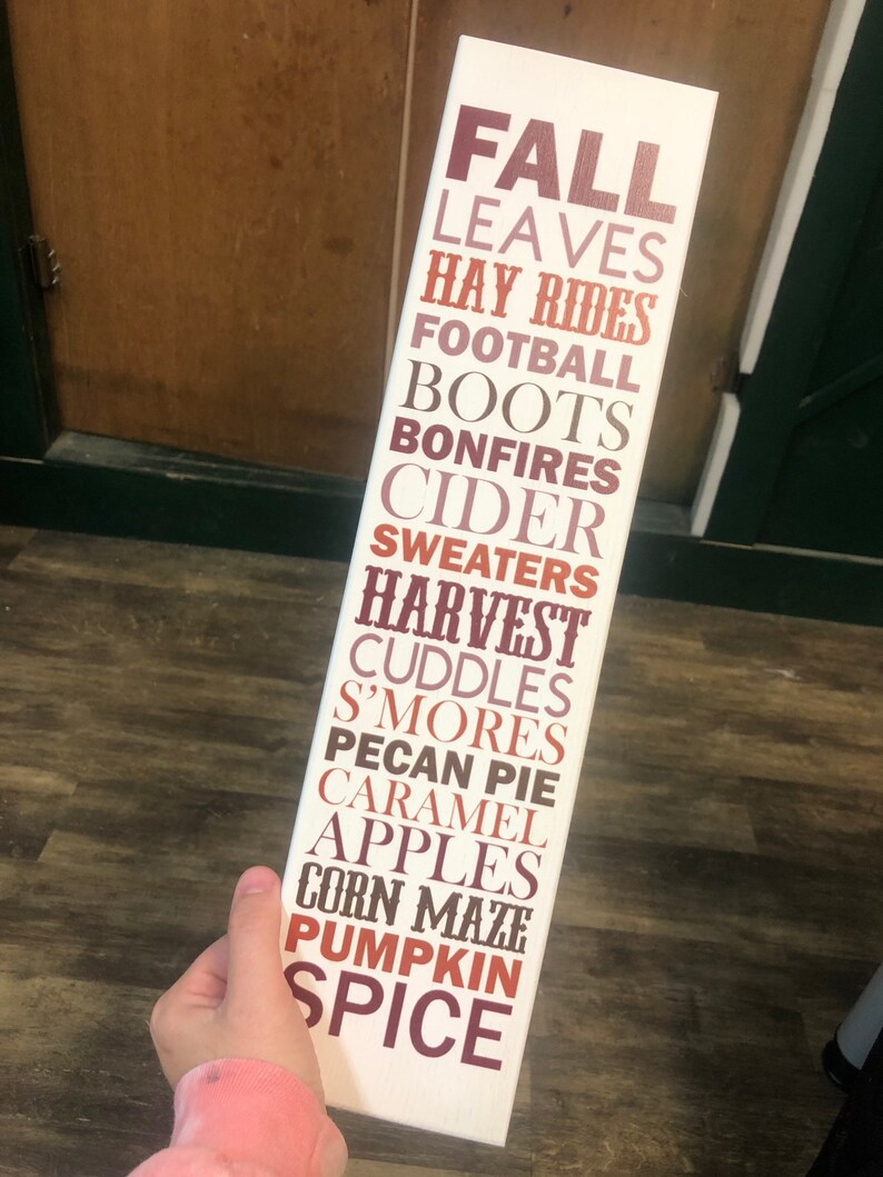 Puede incluir: Un letrero de madera blanco con las palabras "FALL" en letras grandes de color marr&oacute;n. Las palabras "LEAVES", "HAY RIDES", "FOOTBALL", "BOOTS", "BONFIRES", "CIDER", "SWEATERS", "HARVEST", "CUDDLES", "S'MORES", "PECAN PIE", "CARAMEL", "APPLES", "CORN MAZE", "PUMPKIN" y "SPICE" est&aacute;n listadas debajo en letras m&aacute;s peque&ntilde;as, marrones, naranjas y rosadas.