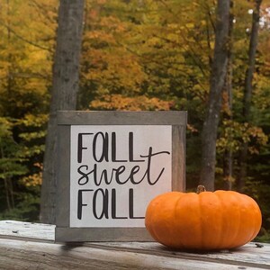May include: A wooden sign with the text "Fall Sweet Fall" in black lettering on a white background. A small orange pumpkin sits in front of the sign.