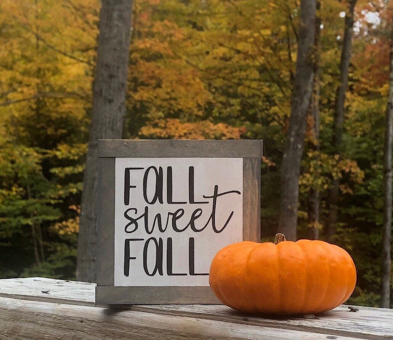 May include: A wooden sign with the text "Fall Sweet Fall" in black lettering on a white background. A small orange pumpkin sits in front of the sign.
