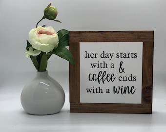 May include: A wooden sign with the text "her day starts with a coffee & ends with a wine" in black lettering on a white background. The sign is framed with a dark brown wood frame.