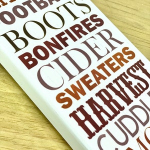 Puede incluir: Un letrero de madera blanco con las palabras "Hay Rides", "Football", "Boots", "Bonfires", "Cider", "Sweaters", "Harvest", "Cuddles" y "Smores" en varios tonos de marr&oacute;n, naranja y gris.