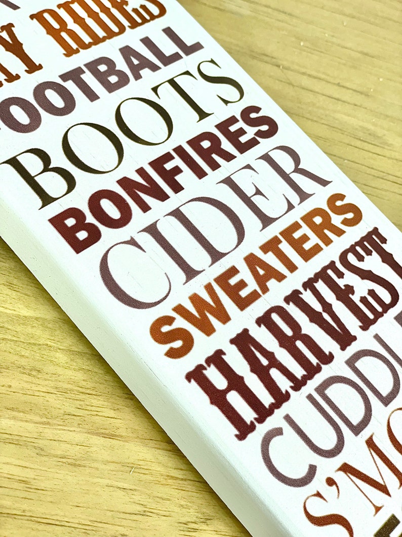 Puede incluir: Un letrero de madera blanco con las palabras "Hay Rides", "Football", "Boots", "Bonfires", "Cider", "Sweaters", "Harvest", "Cuddles" y "Smores" en varios tonos de marr&oacute;n, naranja y gris.