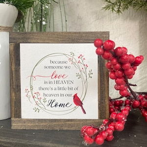 May include: A wooden framed sign with a white background and a green wreath with red berries. The sign reads "because someone we love is in HEAVEN there's a little bit of heaven in our Home". A red cardinal is perched on the wreath.