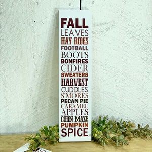 Puede incluir: Un letrero de madera blanco con las palabras "FALL" en letras marrones, seguido de una lista de palabras asociadas con la estaci&oacute;n, incluyendo "leaves", "hay rides", "football", "boots", "bonfires", "cider", "sweaters", "harvest", "cuddles", "s'mores", "pecan pie", "caramel", "apples", "corn maze", "pumpkin", y "spice".