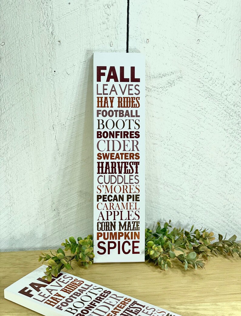 Puede incluir: Un letrero de madera blanco con las palabras "FALL" en letras marrones, seguido de una lista de palabras asociadas con la estaci&oacute;n, incluyendo "leaves", "hay rides", "football", "boots", "bonfires", "cider", "sweaters", "harvest", "cuddles", "s'mores", "pecan pie", "caramel", "apples", "corn maze", "pumpkin", y "spice".