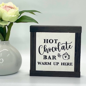 May include: A white wooden sign with black lettering that reads "Hot Chocolate Bar Warm Up Here". The sign has a black frame and a small snowflake and a cup of hot chocolate with whipped cream are depicted next to the word "Bar".