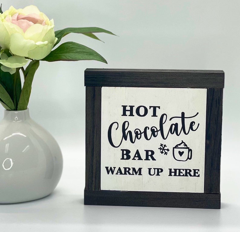 May include: A white wooden sign with black lettering that reads "Hot Chocolate Bar Warm Up Here". The sign has a black frame and a small snowflake and a cup of hot chocolate with whipped cream are depicted next to the word "Bar".