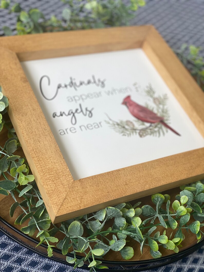 K&ouml;nnte beinhalten: Ein gerahmtes Holzschild mit wei&szlig;em Hintergrund und dem Text "Cardinals appear when angels are near". Ein roter Kardinal sitzt auf einem Zweig mit gr&uuml;nen Bl&auml;ttern und Tannenzapfen.