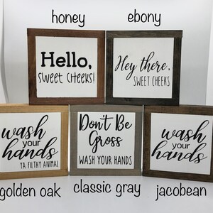 May include: Four wooden signs with different sayings and wood stains. The signs read "Hello, Sweet Cheeks!" in honey stain, "Hey there, Sweet Cheeks" in ebony stain, "Wash your hands, Ya Filthy Animal" in golden oak stain, and "Don't Be Gross, Wash Your Hands" in classic gray stain. The signs are all the same size and shape.