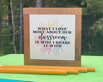 May include: Wooden sign with the text "What I love most about our classroom is who I share it with". The sign has a white background with colorful handprints around the text.