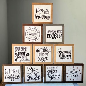 May include: Ten wooden signs with coffee-themed sayings. The signs are arranged in a pyramid shape. The signs have a rustic wood finish and white lettering. The sayings include "Love is brewing", "Fresh brewed coffee served daily", "Life begins after coffee", "Pour some sugar on me - your coffee", "No talkie before coffee", "Caffeine Queen", "But first, coffee", "Rise and grind", "A little bit of coffee and a lotta Jesus", and "Coffee time".