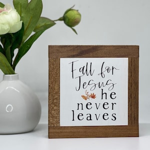 May include: A wooden sign with a white background and the text "Fall for Jesus he never leaves". The sign has a brown wood frame and a small image of fall leaves in the center.