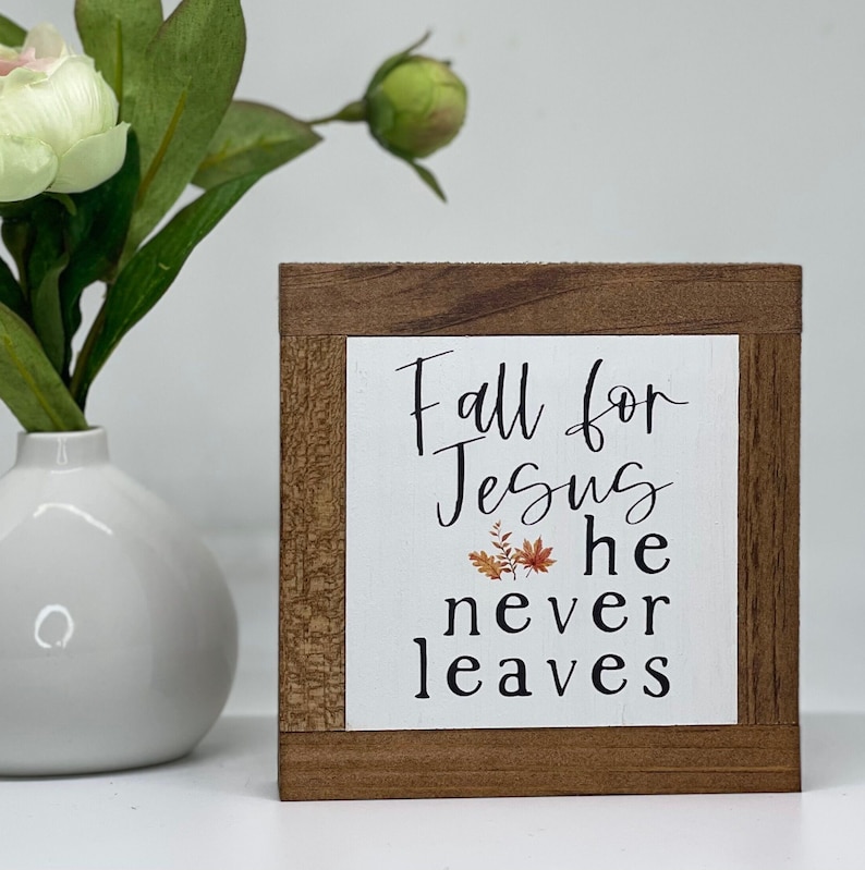 May include: A wooden sign with a white background and the text "Fall for Jesus he never leaves". The sign has a brown wood frame and a small image of fall leaves in the center.