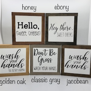 May include: Four wooden signs with different sayings and wood stains. The signs read "Hello, Sweet Cheeks!" in honey stain, "Hey there, Sweet Cheeks" in ebony stain, "Wash your hands, Ya Filthy Animal" in golden oak stain, and "Don't Be Gross, Wash Your Hands" in classic gray stain. The signs are all the same size and shape.