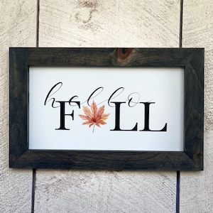 Könnte beinhalten: Rechteckiges Holzschild mit dunkelgrauem Rahmen. Das Schild zeigt die Worte "hello FALL" in einer Mischung aus Schreib- und Blockbuchstaben, mit einem herbstlichen Blatt im Aquarellstil. Der Hintergrund ist weiß.